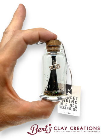 Hand holding a small bottle with a silk graduation tassel inside, labeled '2025', on a white background with 'Bert's Clay Creations' branding.